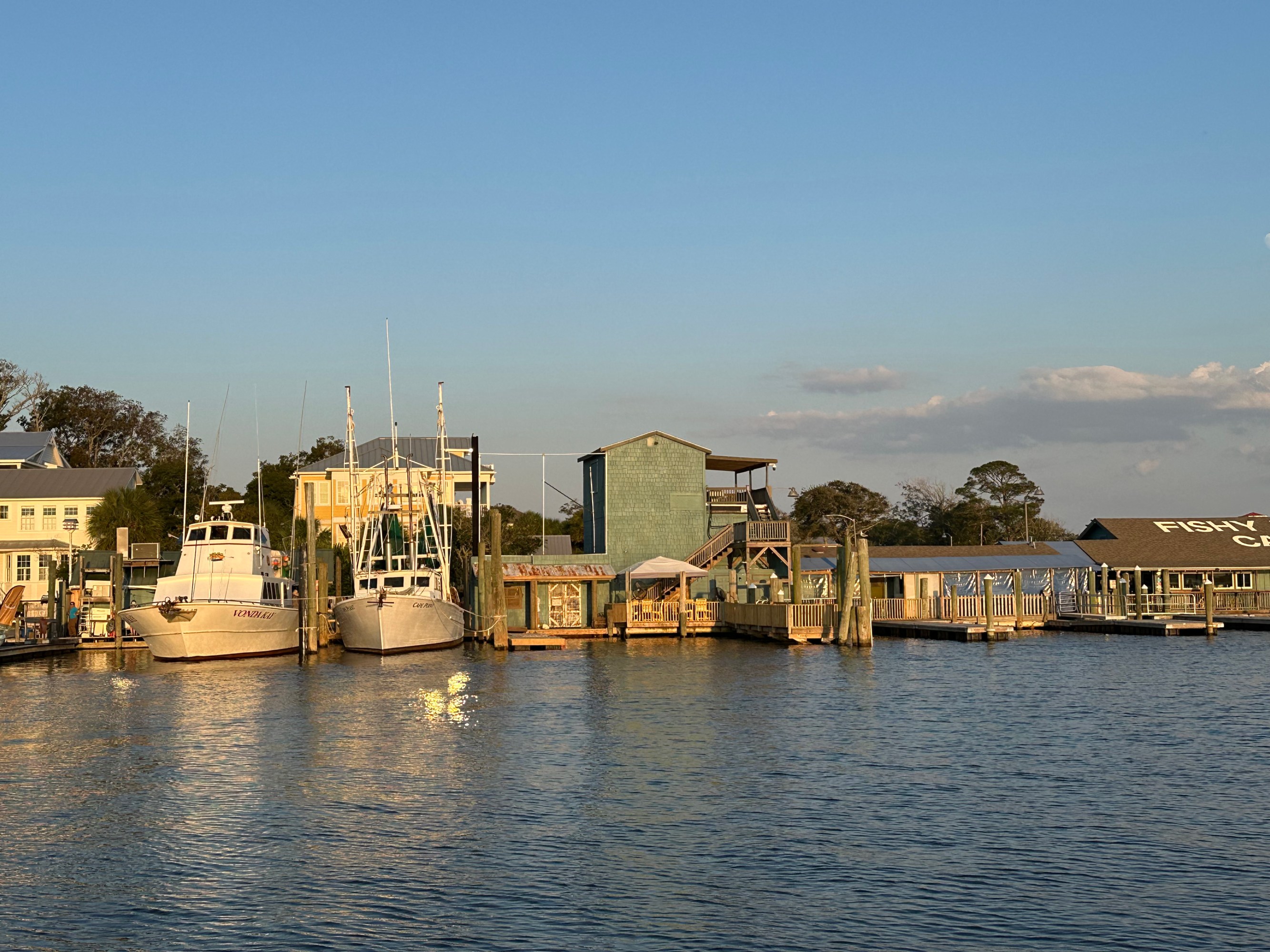 Boats docked at a marina with nearby buildings under a clear blue sky at sunset.